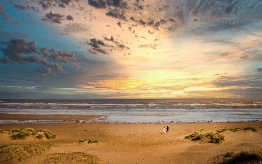 Brudepar går hånd i hånd langs stranden i Vestjylland ved solnedgang – et romantisk bryllupsbillede med en storslået himmel og gyldne klitter.